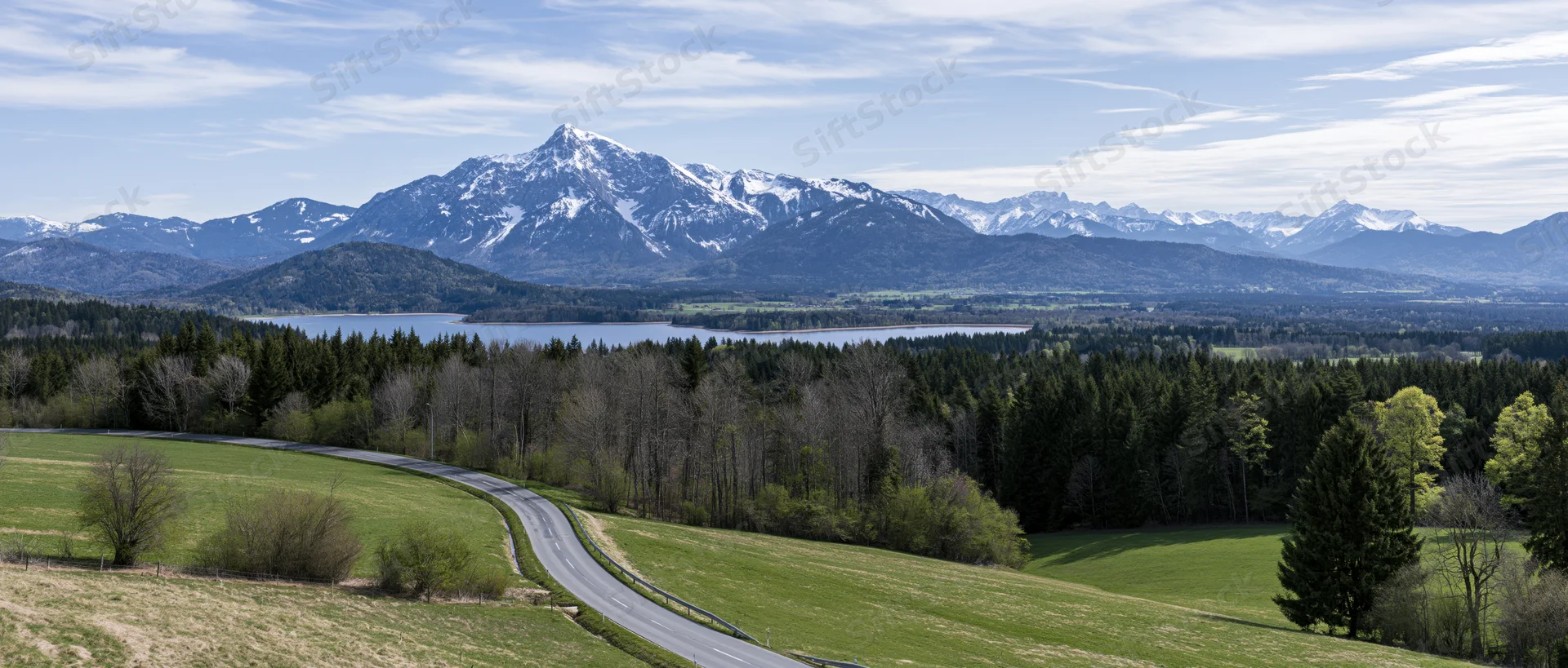 Winding road through green meadows and forest toward snow-capped mountains and a lake in the valley