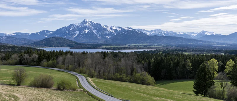 Winding road through green meadows and forest toward snow-capped mountains and a lake in the valley