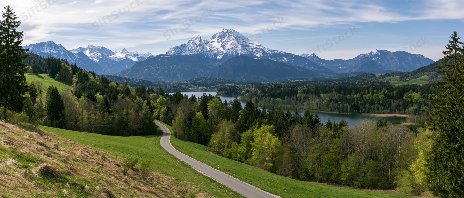 Winding road through green valley with forest and lake leading to snow-capped alpine mountains in the distance
