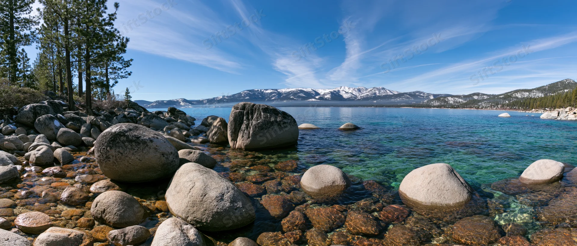 Clear turquoise lake bordered by rocks and pine trees with snow-capped mountains in the distance