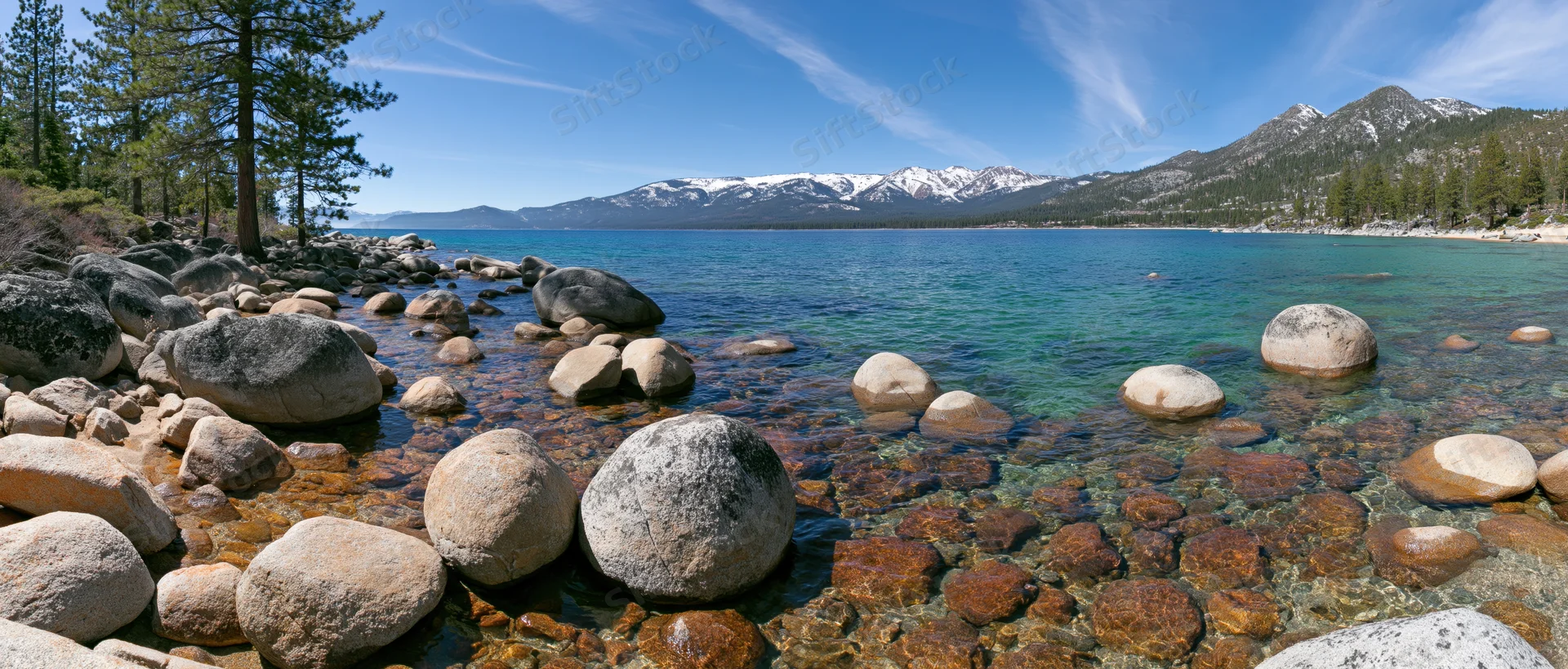 Clear turquoise lake with rocks and pine trees along the shore under blue sky with snow-capped mountains in the distance