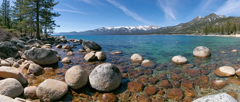 Clear turquoise lake with rocks and pine trees along the shore under blue sky with snow-capped mountains in the distance