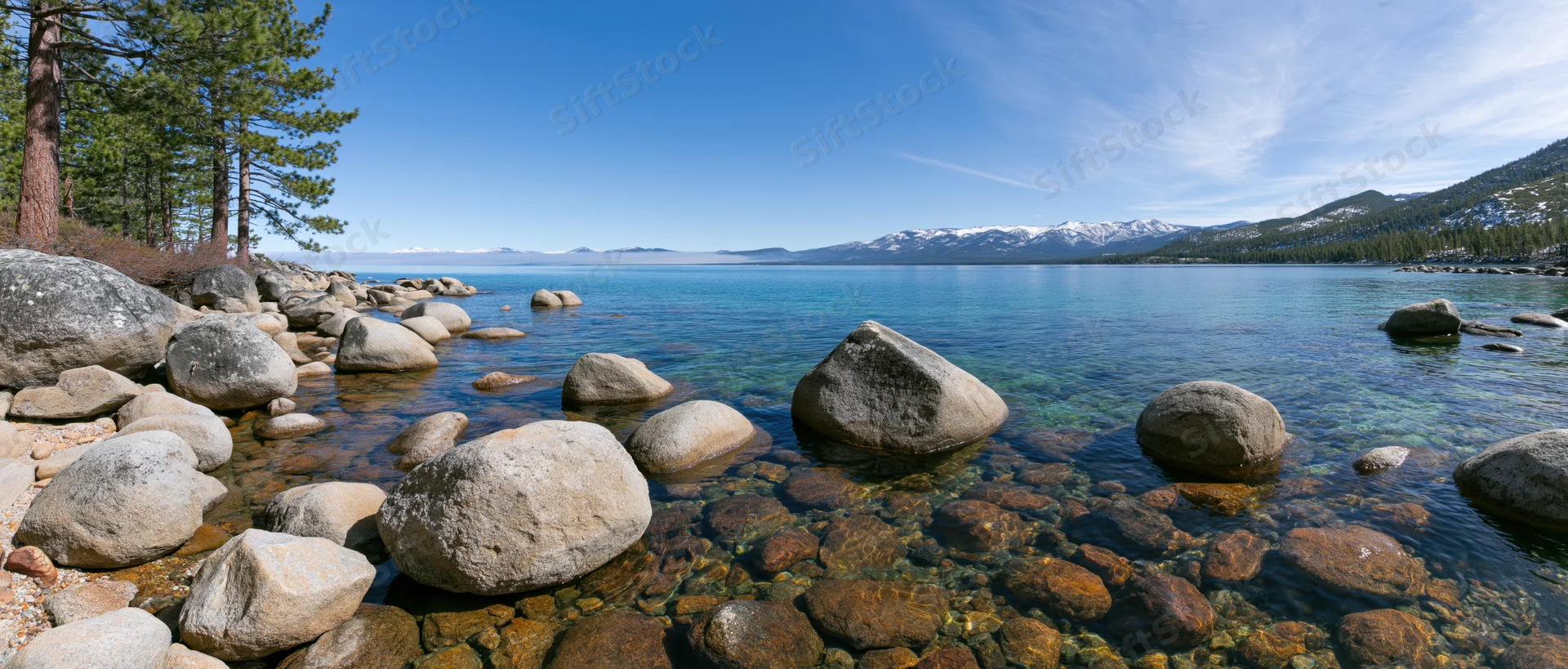 Clear turquoise lake with rocks along the shore and snow-capped mountains in the distance under a blue sky