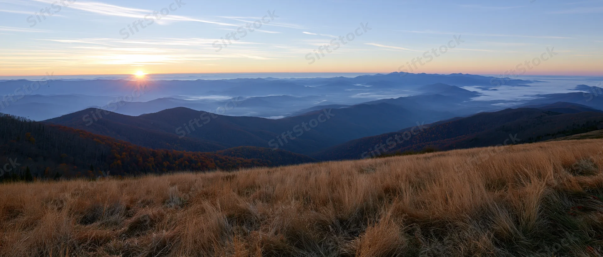 Golden sunrise over layered mountain ranges with autumn forest and grassy meadow in foreground