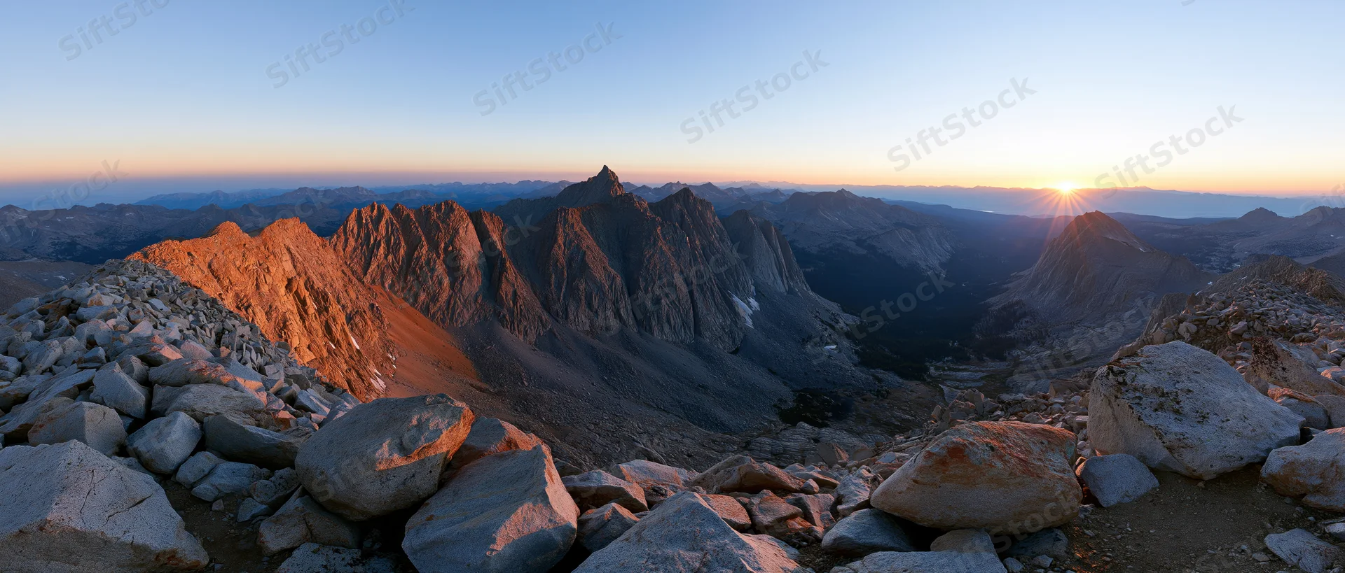 Rocky mountain peaks at sunrise overlooking a valley with boulders in the foreground