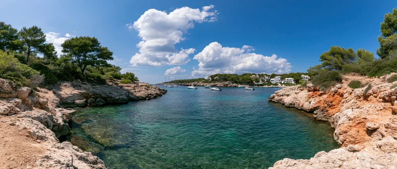 Turquoise water in a rocky cove with boats and white villas along the shoreline under a blue sky with clouds