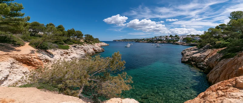 Turquoise bay surrounded by rocky cliffs and pine trees with white houses and boats in the distance