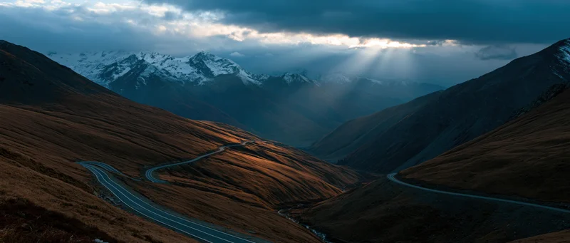 Winding asphalt road through alpine valley with snow-capped peaks under cloudy sky at dusk