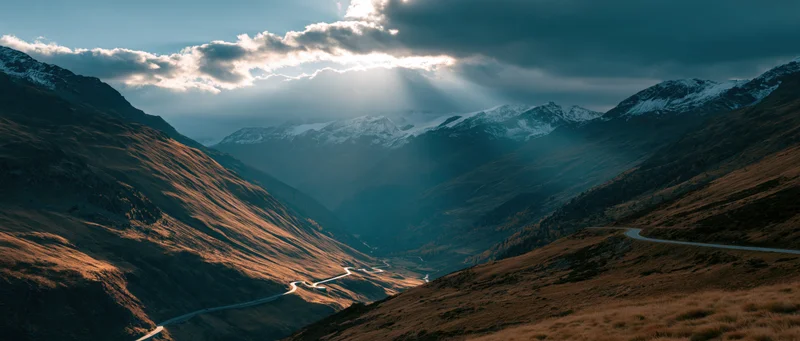 Winding road through mountain valley at sunset with snow-capped peaks and sunbeams through clouds