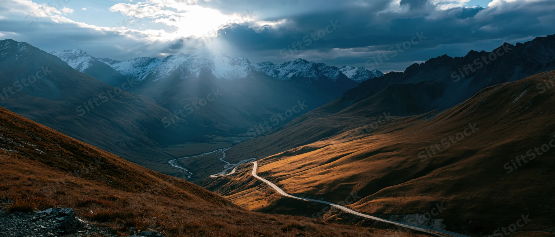 Winding road through alpine valley with snow-capped peaks and golden slopes under dramatic sky