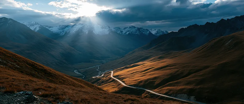 Winding road through alpine valley with snow-capped peaks and golden slopes under dramatic sky