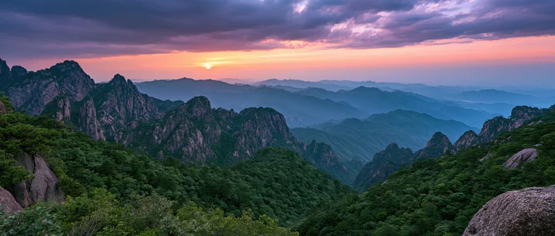 Mountain range at sunset with layered peaks and forested valleys under colorful twilight sky