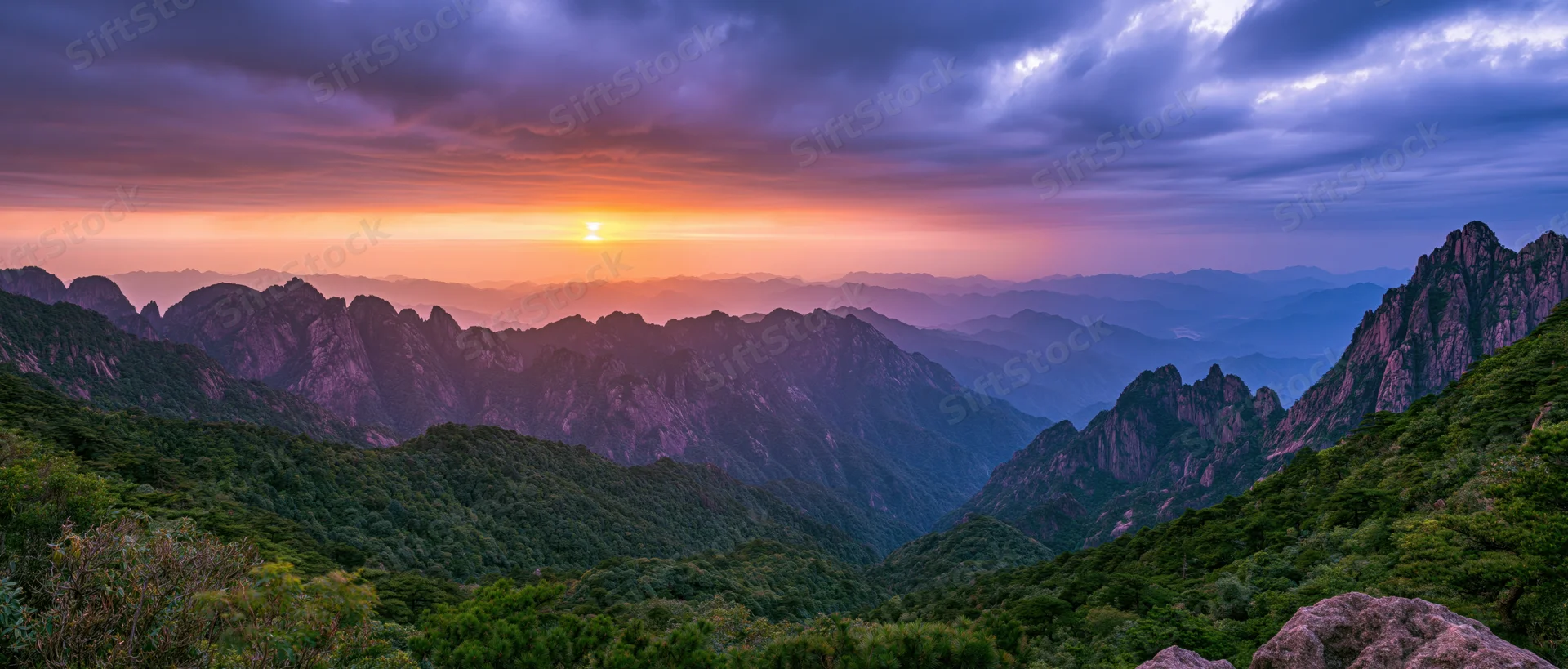 Mountain range at sunset with layered peaks and forested valleys under colorful sky