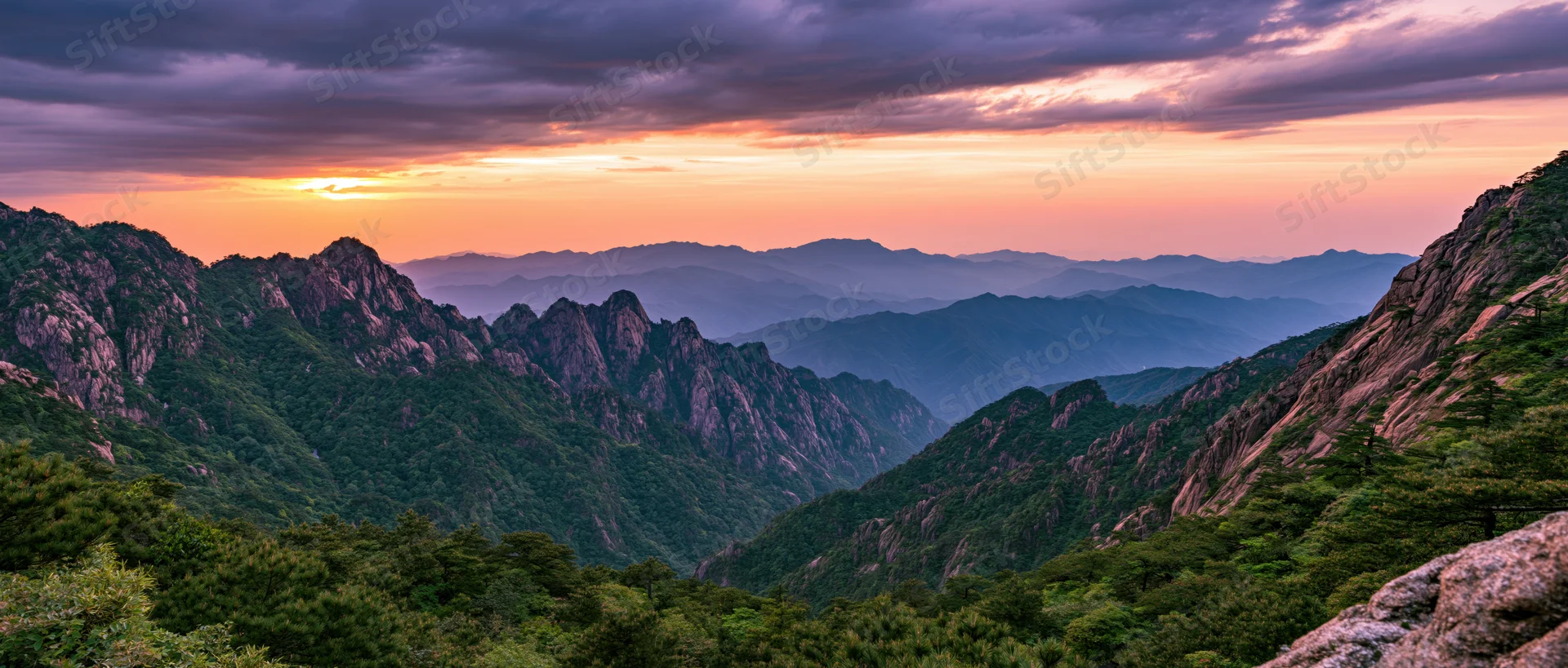 Mountain range at sunset with layered ridges and forested valleys under colorful sky