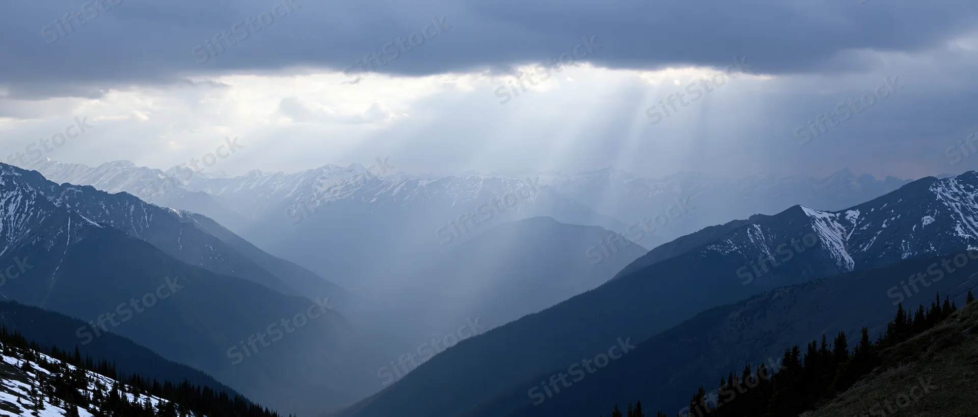 Snow-covered mountain range with sun rays breaking through clouds over a deep valley