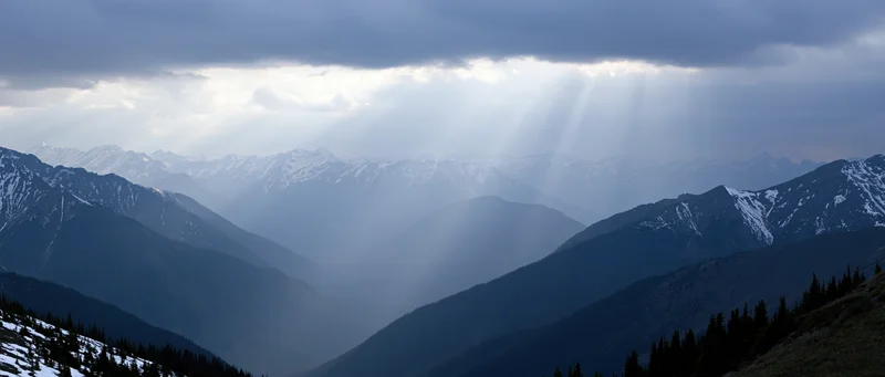 Snow-covered mountain range with sun rays breaking through clouds over a deep valley