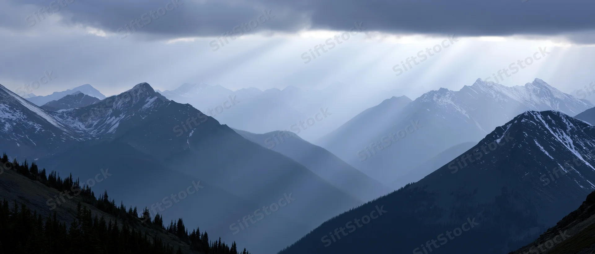 Snow-capped mountain range with crepuscular rays breaking through clouds over a misty valley lined with evergreen trees