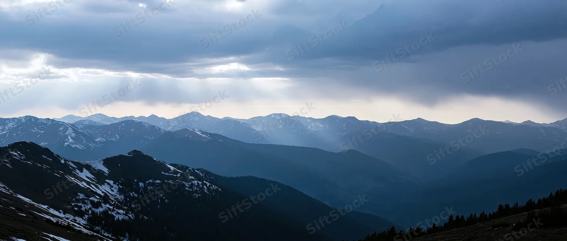 Snow-dusted mountain range under stormy sky with sunbeams breaking through clouds