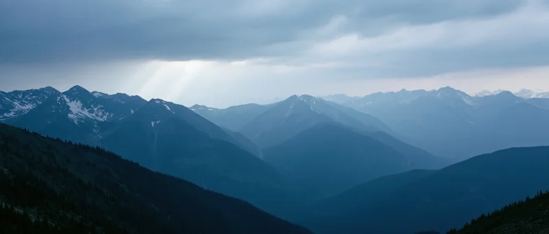 Layered mountain range with snow-capped peaks and forested slopes under cloudy sky