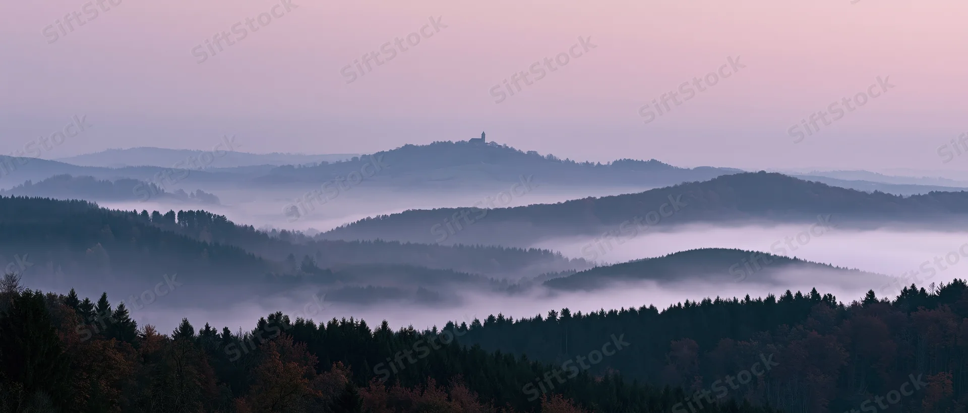 Misty forested hills under soft dawn light with distant tower on ridge
