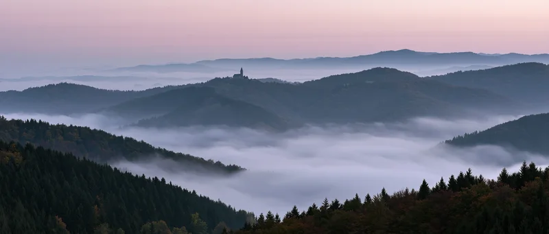 Misty valley with forested hills and distant castle tower at dawn