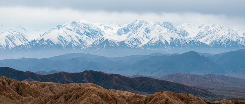 Layered mountain range with snow-capped peaks and arid foothills under overcast sky