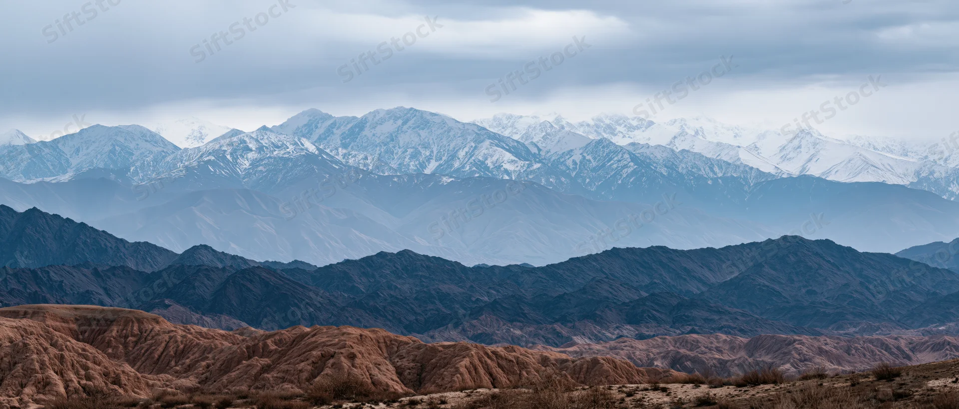 Snow-capped mountain range layered with arid desert terrain under overcast sky