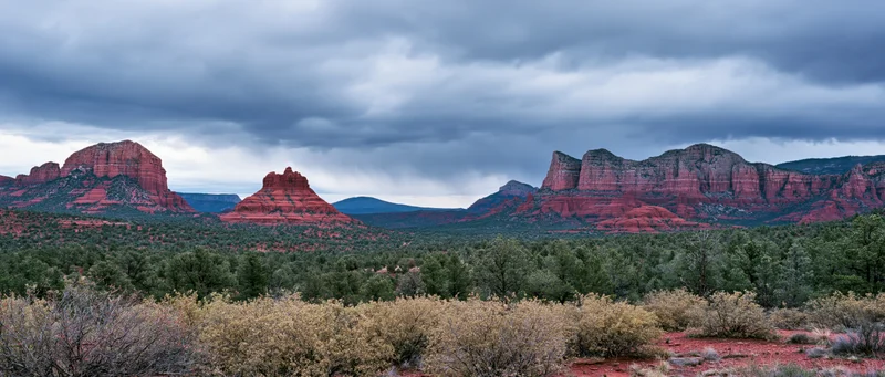 Red rock formations rising from desert valley with juniper forest and shrubs under cloudy sky