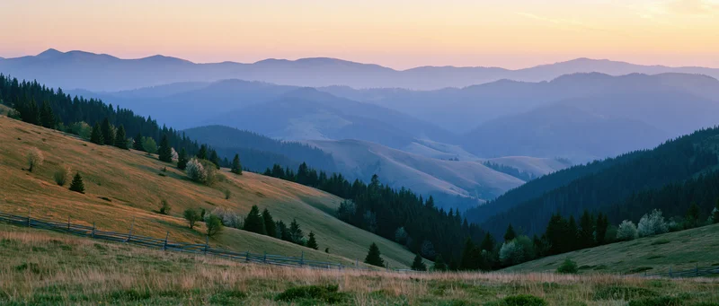 Rolling hills and forested valleys under a hazy sunset sky with distant mountain ranges and a wooden fence in the foreground
