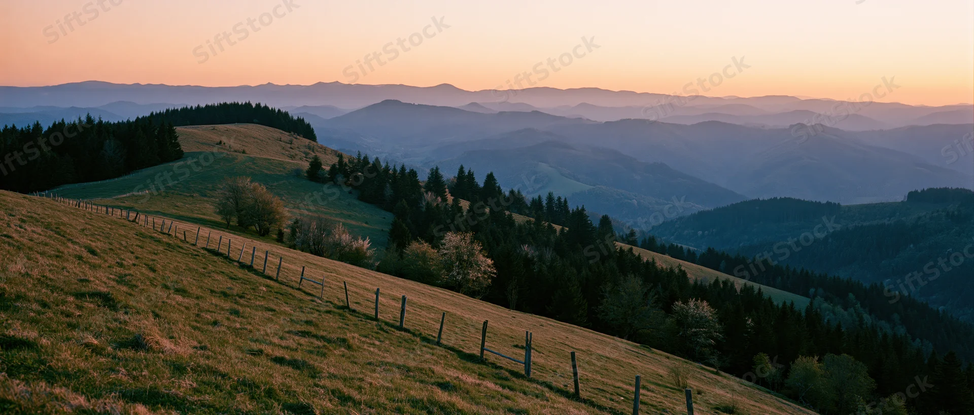 Rolling hills covered in grass and trees with a wooden fence, overlooking a mountain valley at sunset