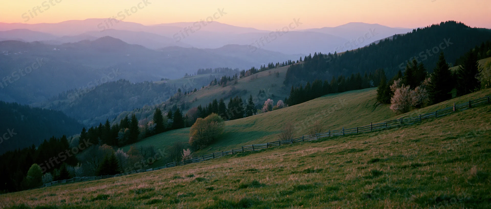 Rolling hills covered in grass and trees with a wooden fence under a sunset sky