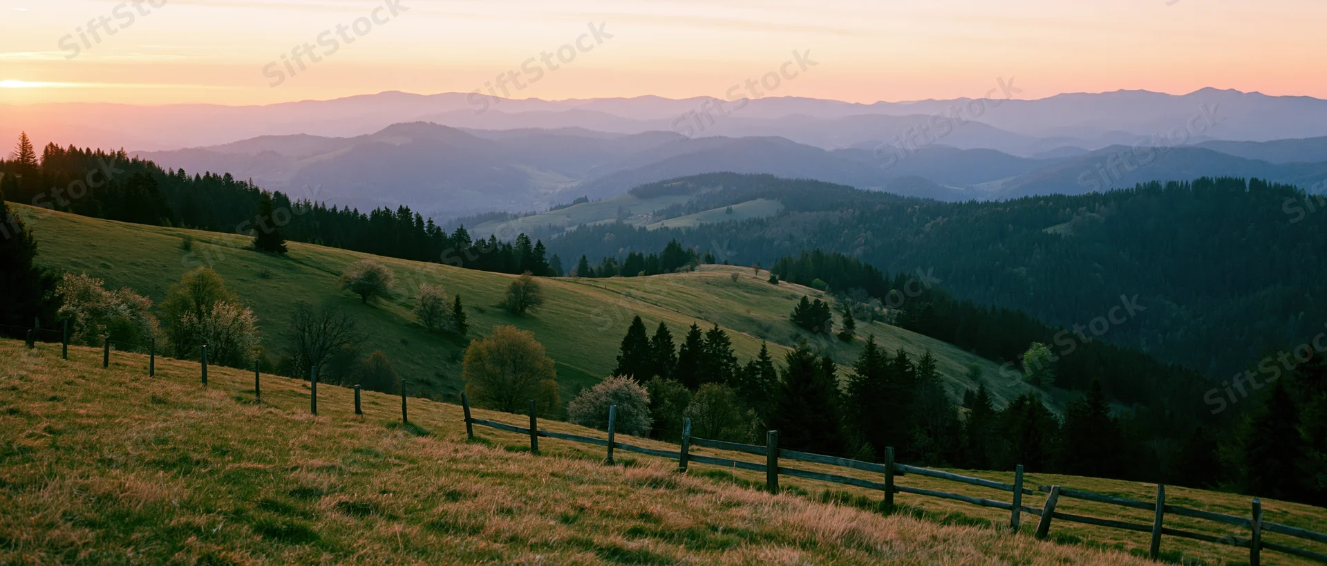 Rolling hills covered in grass and trees with a wooden fence in the foreground under a sunset sky
