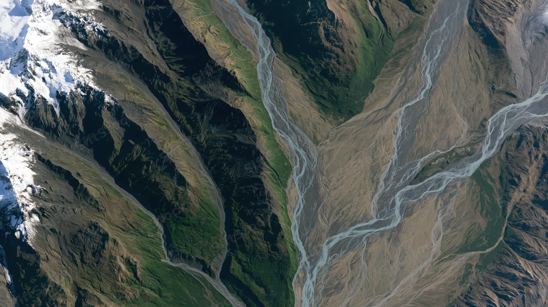 Aerial view of a mountain valley with snow-capped peaks, rivers flowing through rugged terrain and green slopes