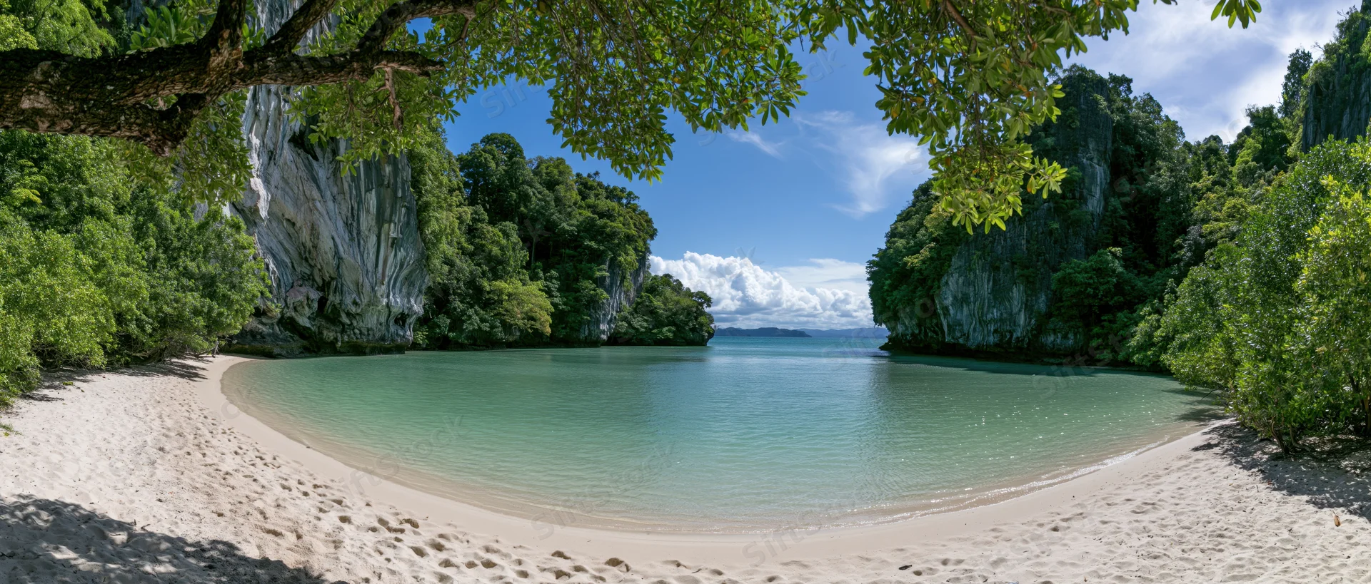 Curved sandy beach surrounded by lush green trees and limestone cliffs opening to a calm turquoise lagoon under a blue sky with scattered clouds