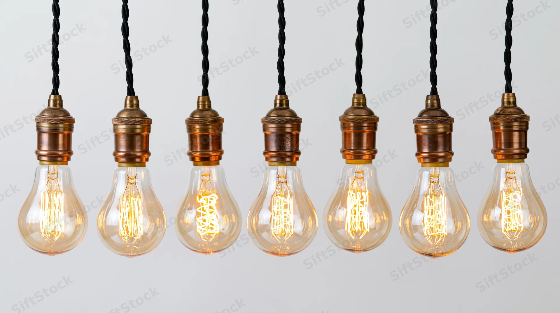 Seven vintage Edison lightbulbs hanging in a row with copper bases and black twisted cords against a plain background