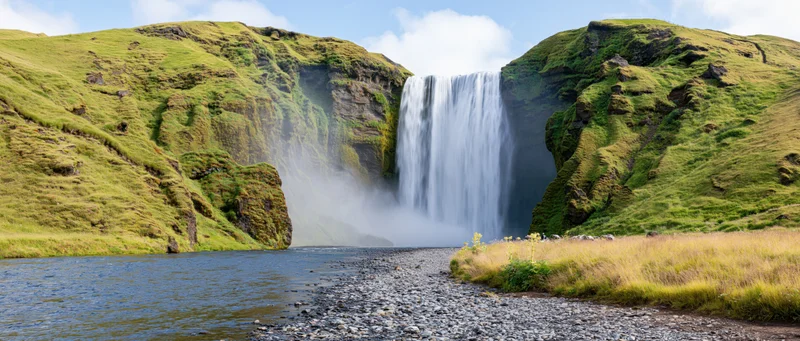 Waterfall cascading down moss-covered cliffs into a river surrounded by green hills and grassy meadows