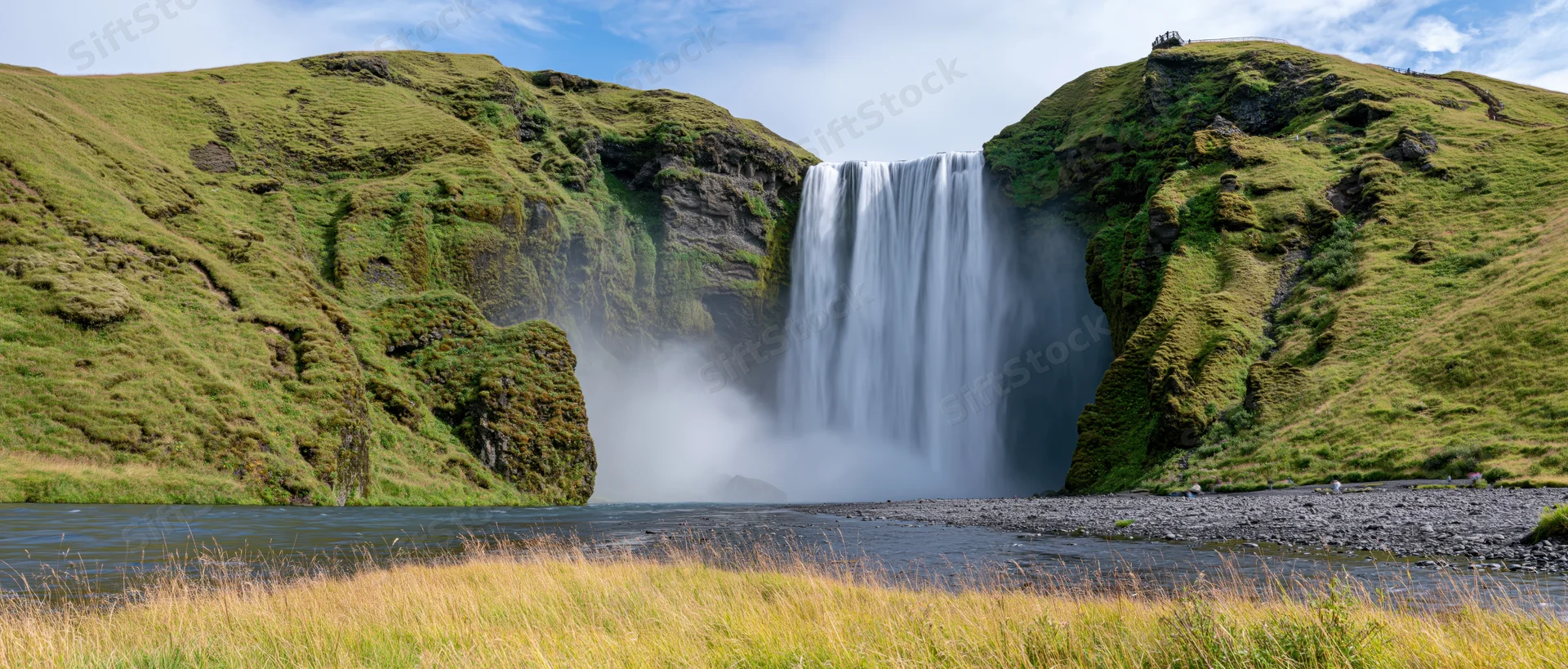 Wide view of a powerful waterfall cascading between moss-covered cliffs into a river below in a green valley