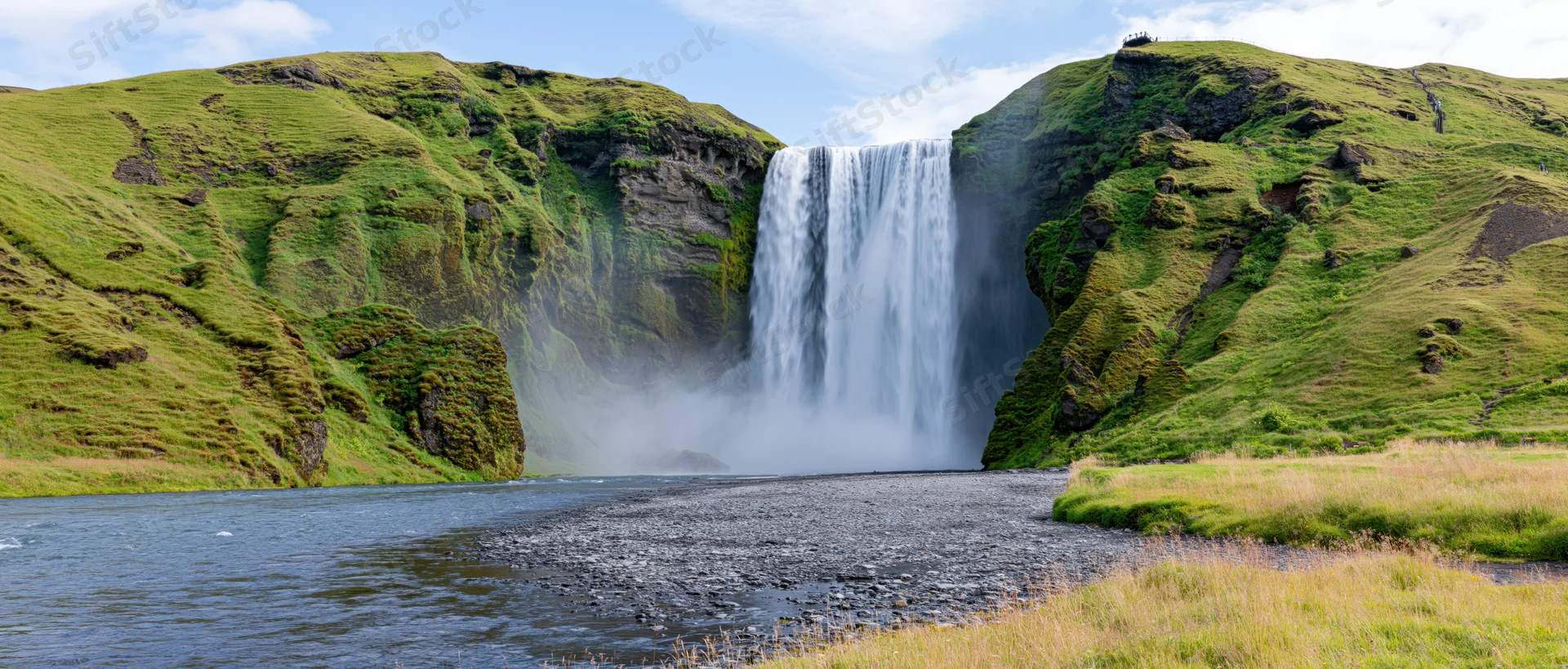 Wide waterfall cascading down moss-covered cliffs into a river surrounded by green hills under a blue sky