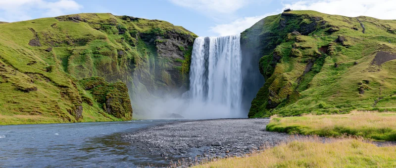 Wide waterfall cascading down moss-covered cliffs into a river surrounded by green hills under a blue sky