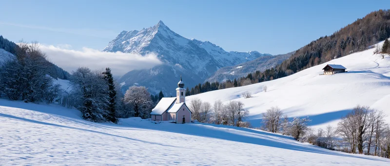 Small white chapel with steeple on snow-covered hillside in alpine valley with snow-capped mountain in background