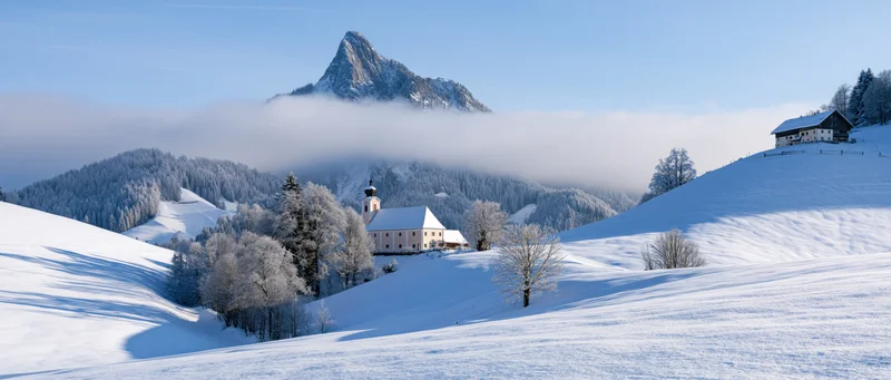 White church nestled in snow-covered alpine valley with fog-shrouded mountain peak in background