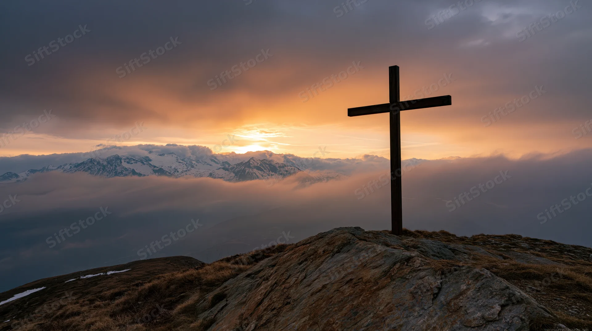 Dark wooden cross silhouetted on rocky mountain peak at sunset with snow-capped peaks and clouds below