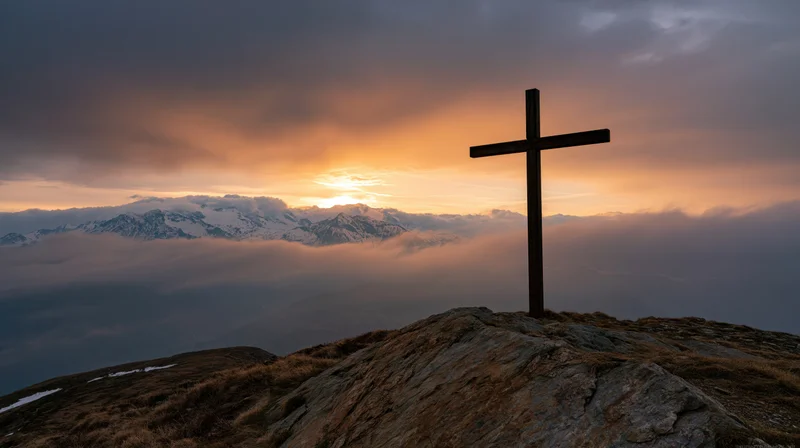 Dark wooden cross silhouetted on rocky mountain peak at sunset with snow-capped peaks and clouds below