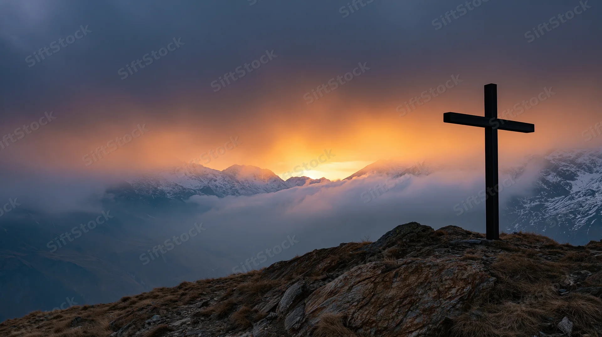 Dark wooden cross silhouetted on rocky mountain peak at sunset with snow-capped peaks and clouds below