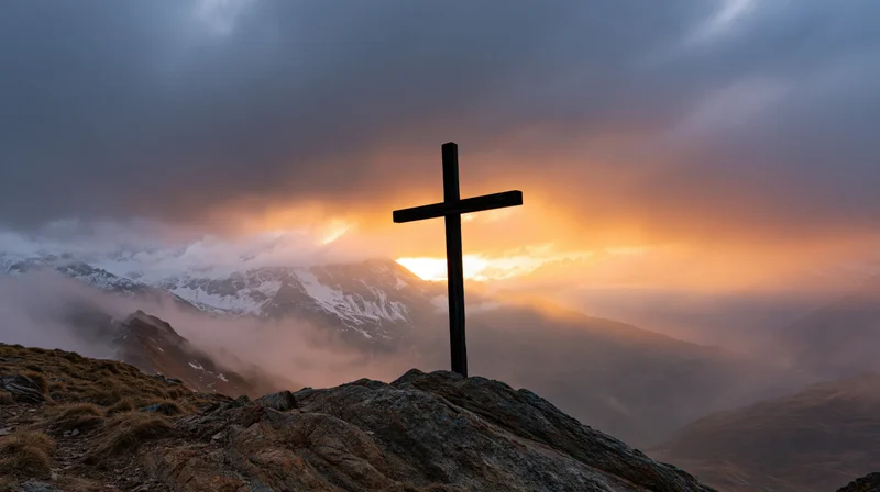 Dark wooden cross silhouetted on rocky mountain peak at sunset with snow-capped peaks and clouds in background