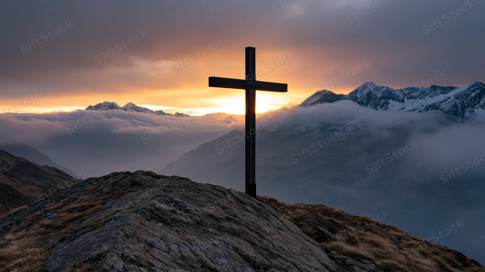 Dark wooden cross silhouetted on rocky mountain summit at sunset above clouds