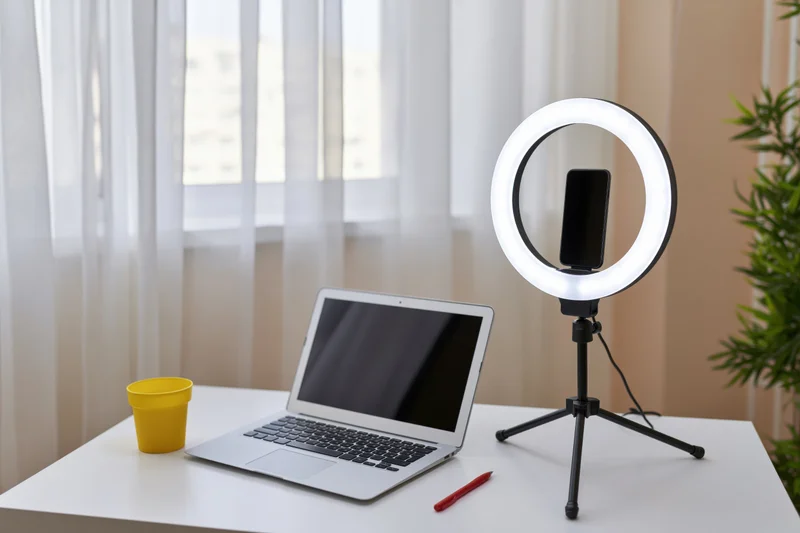 Open laptop and ringlight with smartphone on tripod on white desk near window with plant and yellow cup