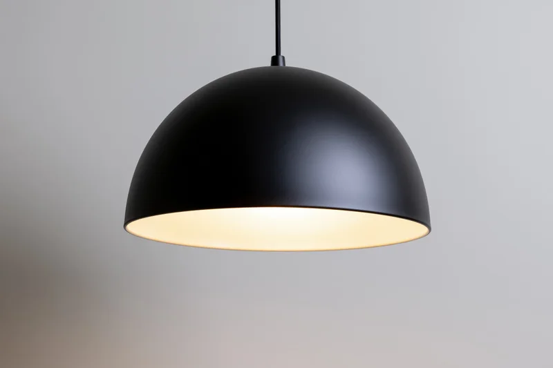 Matte black dome pendant lamp hanging from ceiling with warm glow against plain background