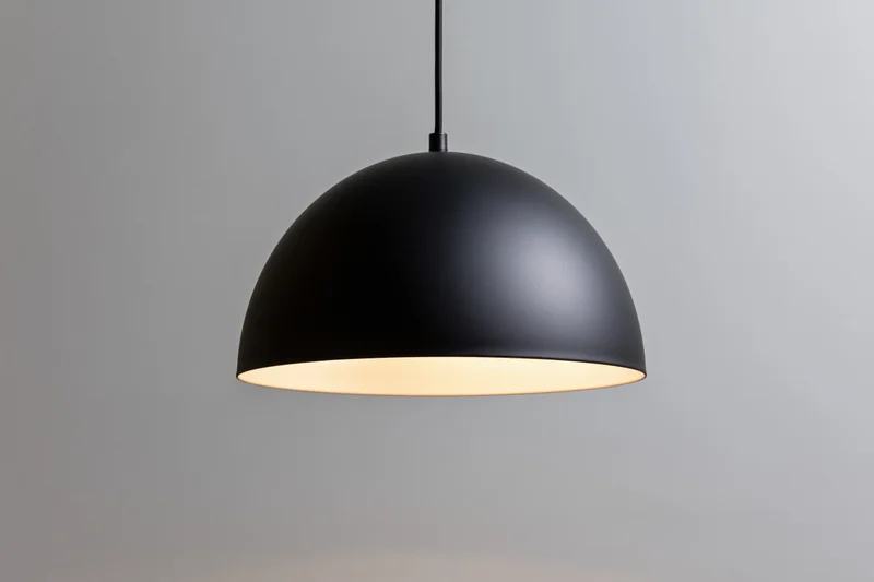 Matte black dome pendant lamp hanging from ceiling with warm glow against plain background