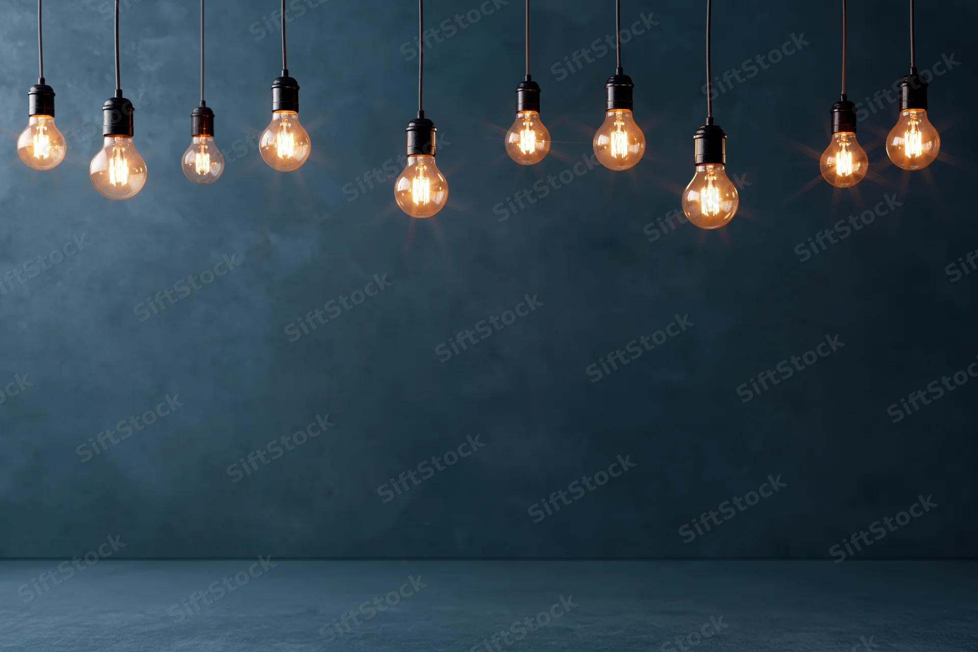 Row of glowing incandescent bulbs hanging from cords against a dark blue concrete wall with copyspace below
